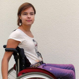 Foto: Akina sitzt wieder im Shakira-T-Shirt und einer lila Jeans sehr souverän in ihrem Rolli vor einer weißen Wand.