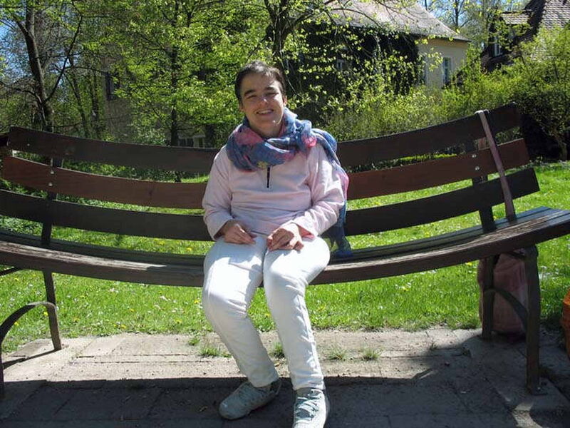 Foto: Die lächelnde Uschi sitzt in heller Kleidung auf einer absichtlich gebogenen, lächelnden, braunen Bank.