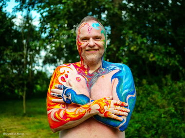 Ich stehe mit bunt bemalten Armen mit den Themen "Feuer" rechts und "Wasser" links in einem grünen Garten.