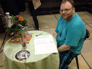 Foto: Ich sitze mnit hellblauem T-Shirt und meiner hell- und dunkelblauen Wickelhose an meinem Lesetisch mit Blumenstrauß.