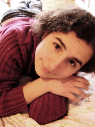 Foto: Mio schaut auf ihrem Bett liegend in die Kamera. Dabei stützt sie den Kopf auf den rechten Handrücken.