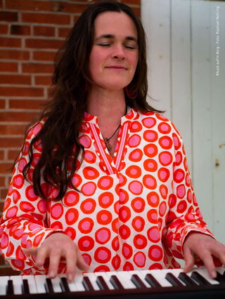 Marie-Lou sitzt mit einer Bluse mit rosaroten Kringeln an einem Piano und spielt Musik.